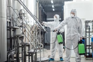 Sanitation at factory of craft beer. Guys in protective suits disinfect at plant