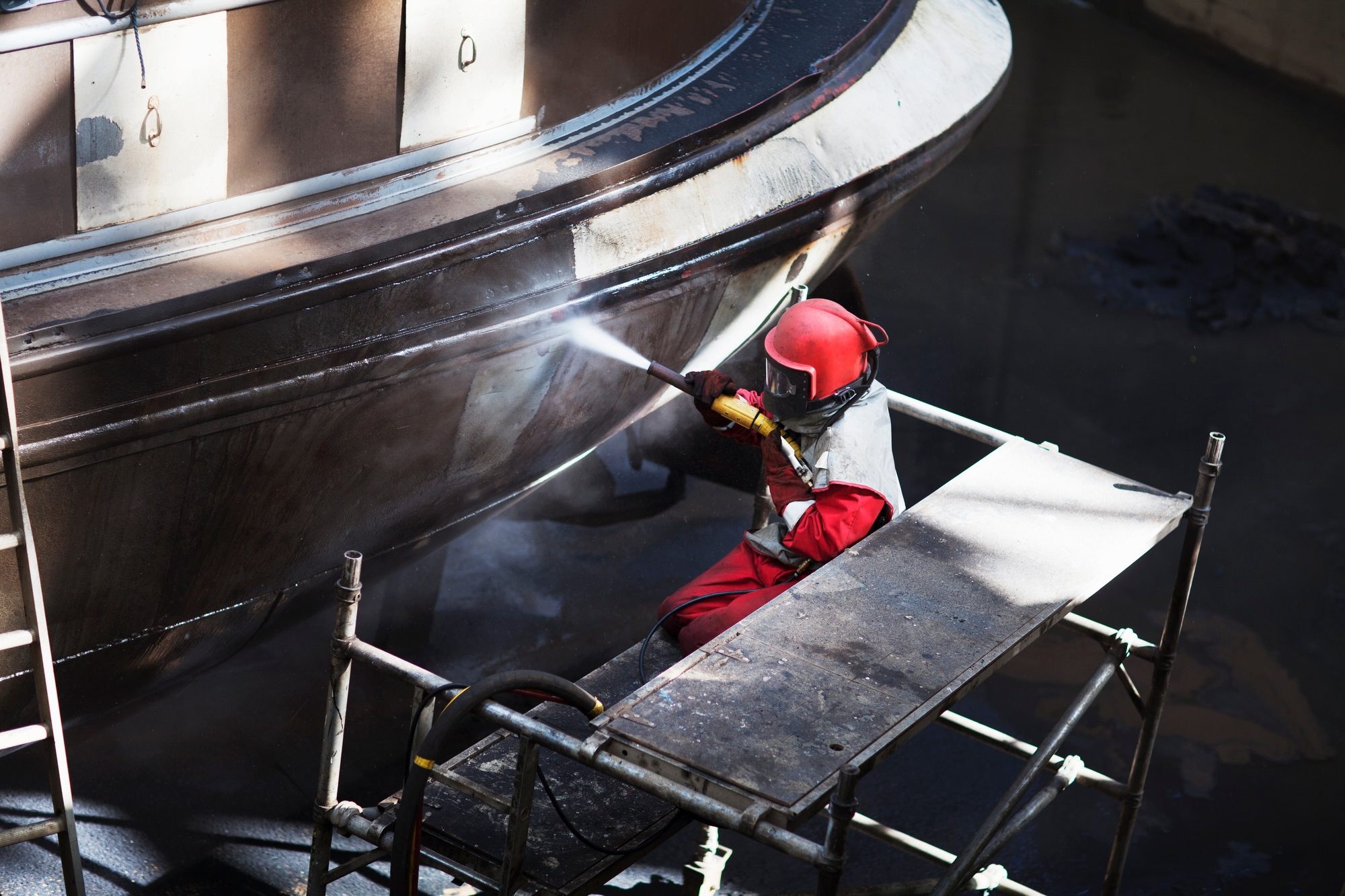 Worker cleaning hull of boat with high pressure hose in shipyard